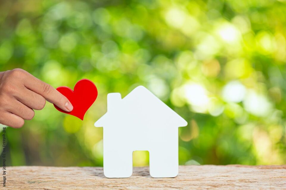 Bild zeigt ein weißes, stilisiertes Hausmodell vor einem grünen Hintergrund, von links kommt eine Hand ins Bild die ein rotes Herz in die Nähe des Hauses hält - Blogartikel warum-es-so-toll-ist-ein-eigenes-haus-zu-bauen-blog-frau-fertighaus