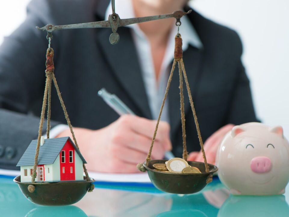 Symbolfoto für Immobilienbewertung, Bild zeigt eine altmodische Waage mit zwei Schalen, in einer liegt ein Modellhaus, in der anderen liegen Münzen, daneben ein Sparschwein, im Hintergrund schreibt eine Person etwas auf, Blog Frau Fertighaus