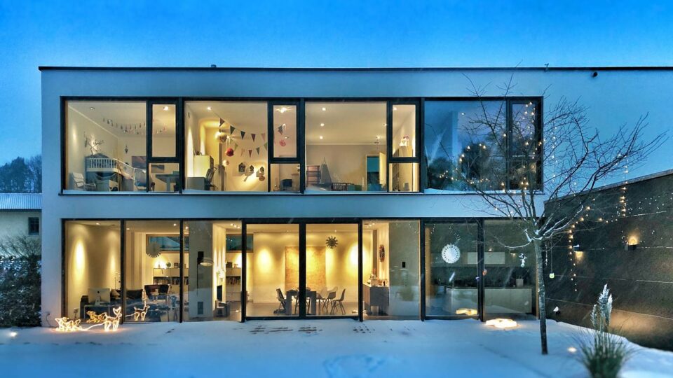 Bild zeigt ein modernes Haus mit Flachdach, große Fensterfronten, Haus hell erlauchtet in der Dämmerung, Winter, Schnee, Hausbeispiel Frau Fertighaus Webseite