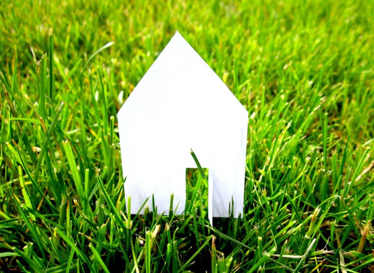 Bild zeigt ein weißes Hausmodell aus Papier auf einer grünen Wiese, Beitragsbild Blogartikel warum-es-so-toll-ist-ein-eigenes-haus-zu-bauen-01-blog-frau-fertighaus
